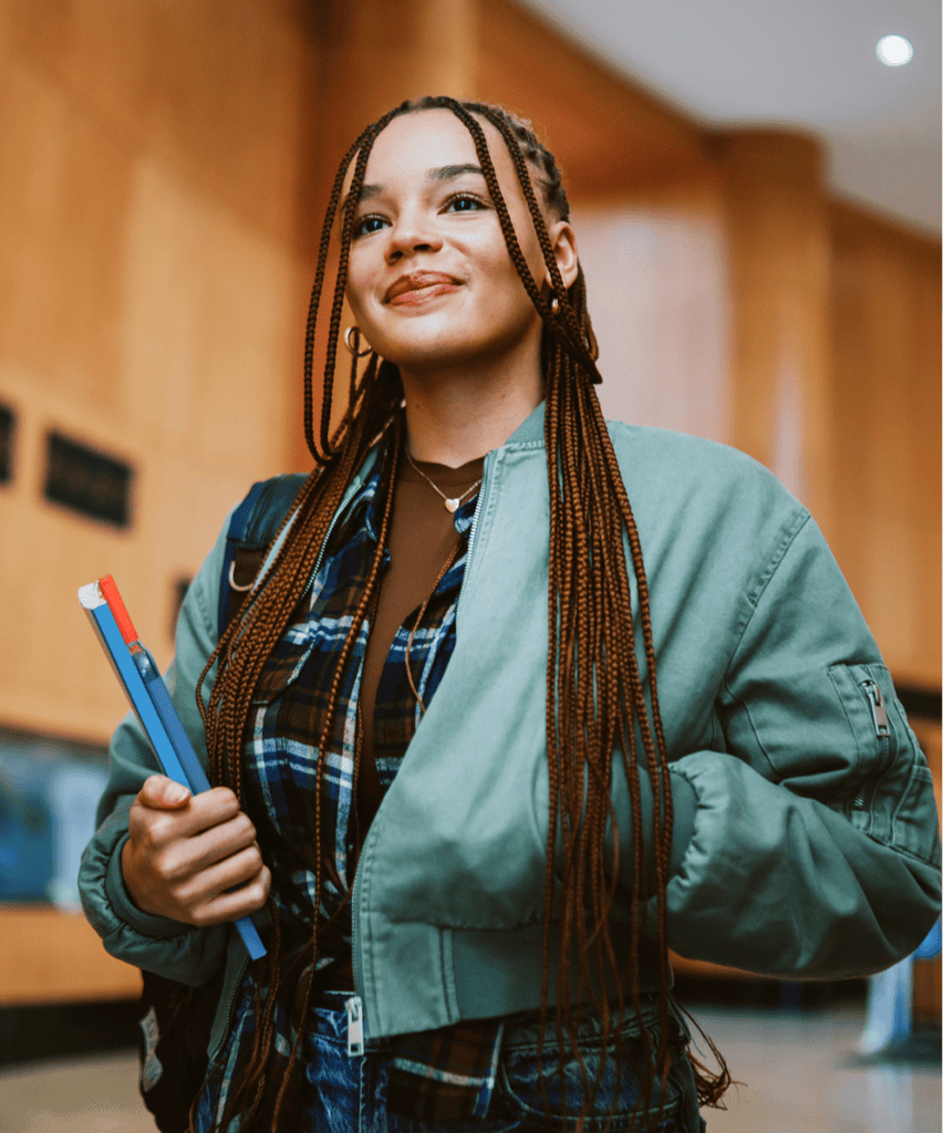 Young woman with braided hair holding a blue pencil, wearing a sage green jacket and plaid shirt.