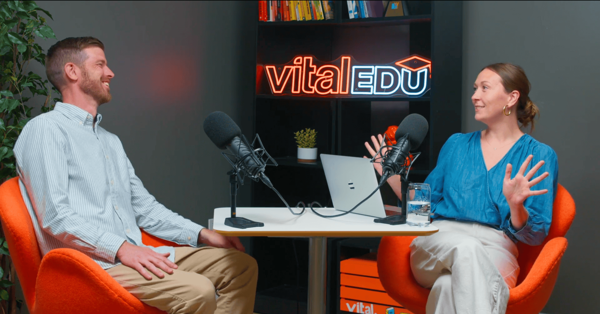 A man and a woman sit in on either side of a table with a pair of microphones on it. A neon sign that says "vitalEDU" is on a shelf behind them. The man is laughing as the woman talks animatedly.